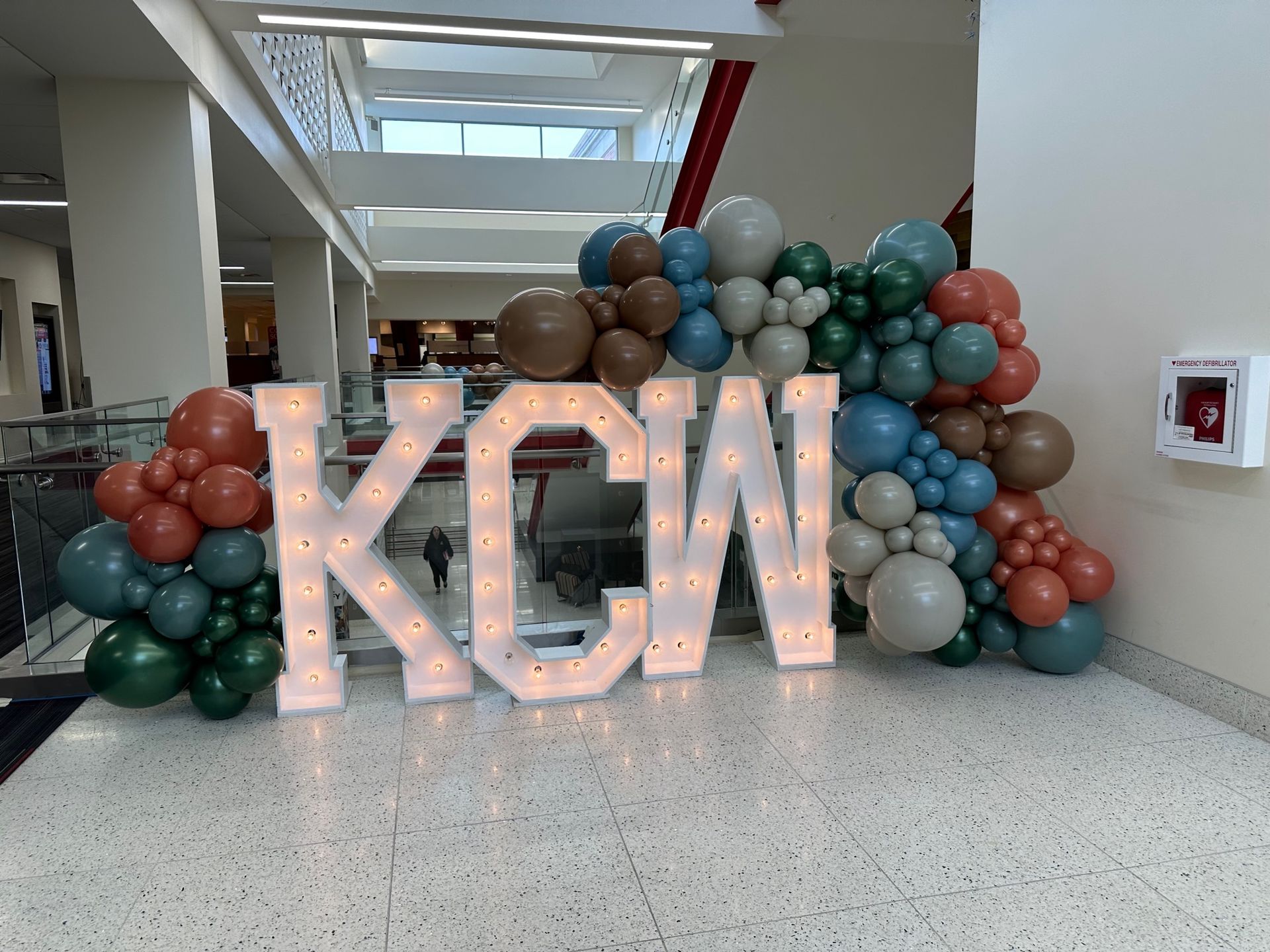 A large lighted letter k is surrounded by balloons in a hallway.