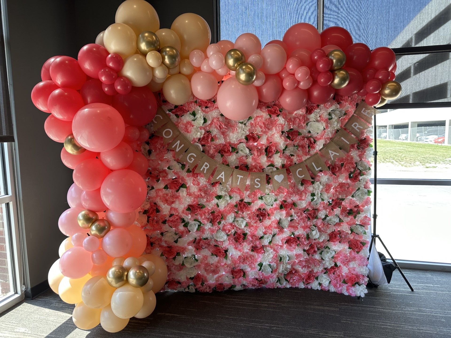 A wall of balloons and flowers is sitting in front of a window.