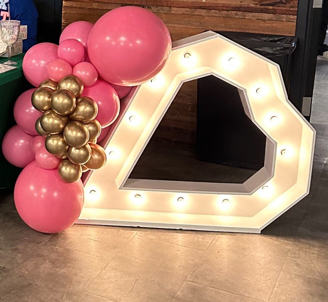 A large heart shaped lighted sign is surrounded by pink and gold balloons.