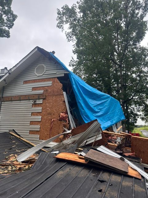Damaged house roof with tarp and debris after storm, collapsed siding and scattered debris outside.