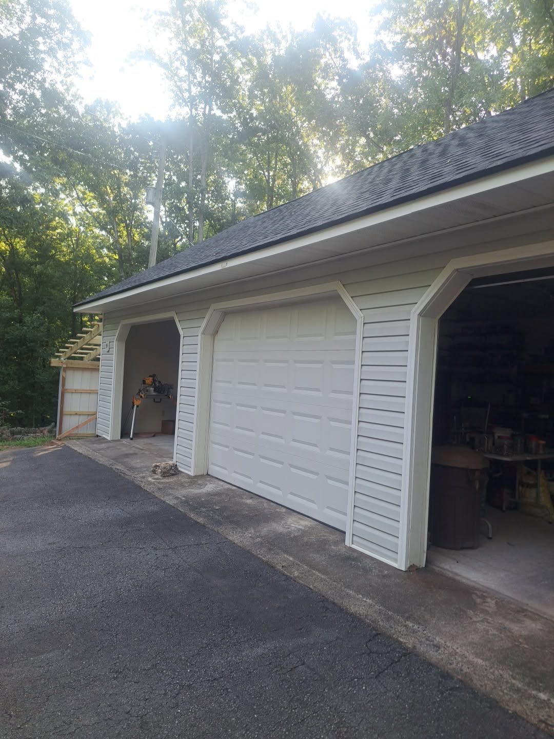 Two-car garage with open bays, white siding, and a paved driveway under trees.