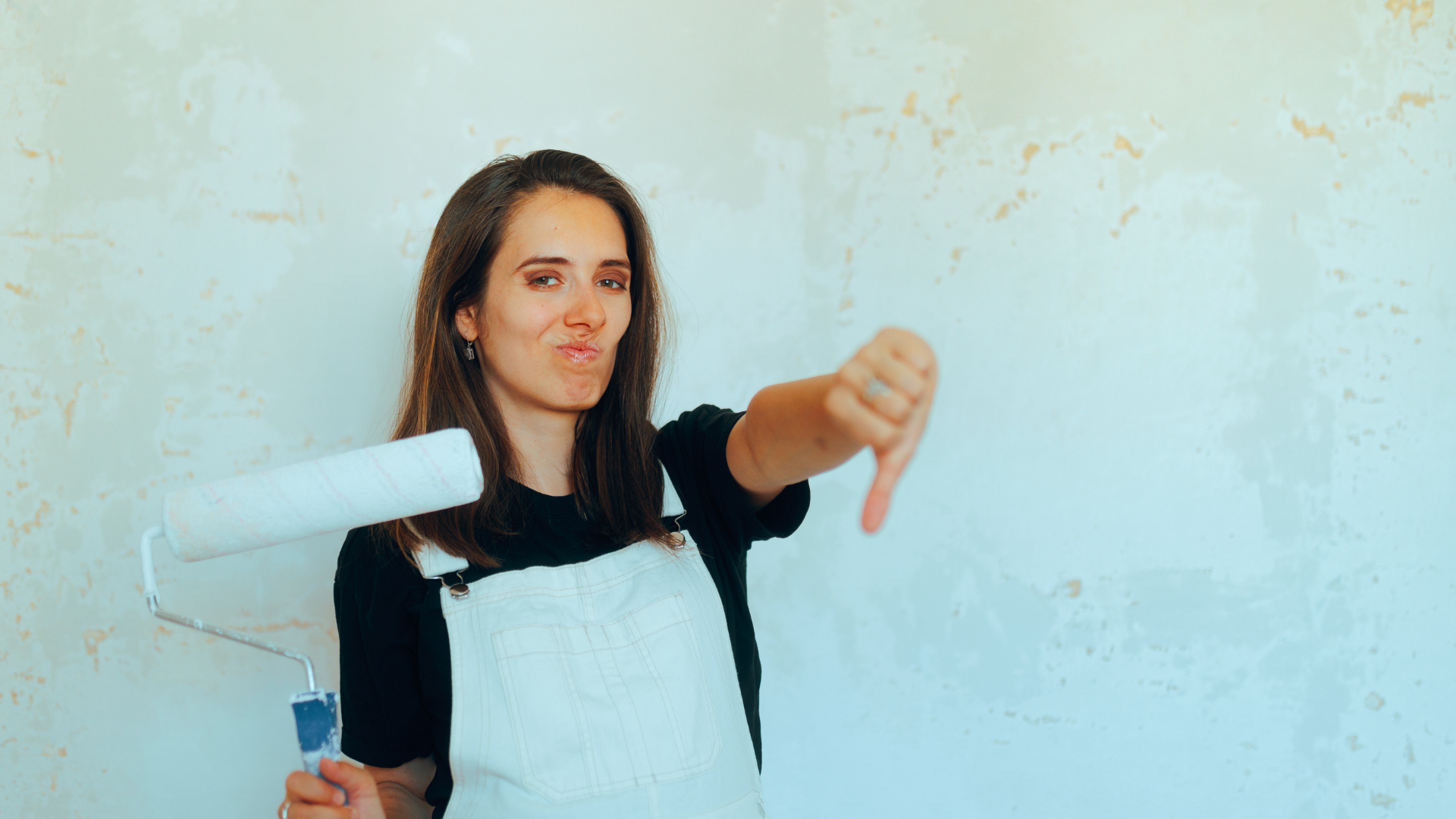 Woman with paint roller gives thumbs down in front of a wall.