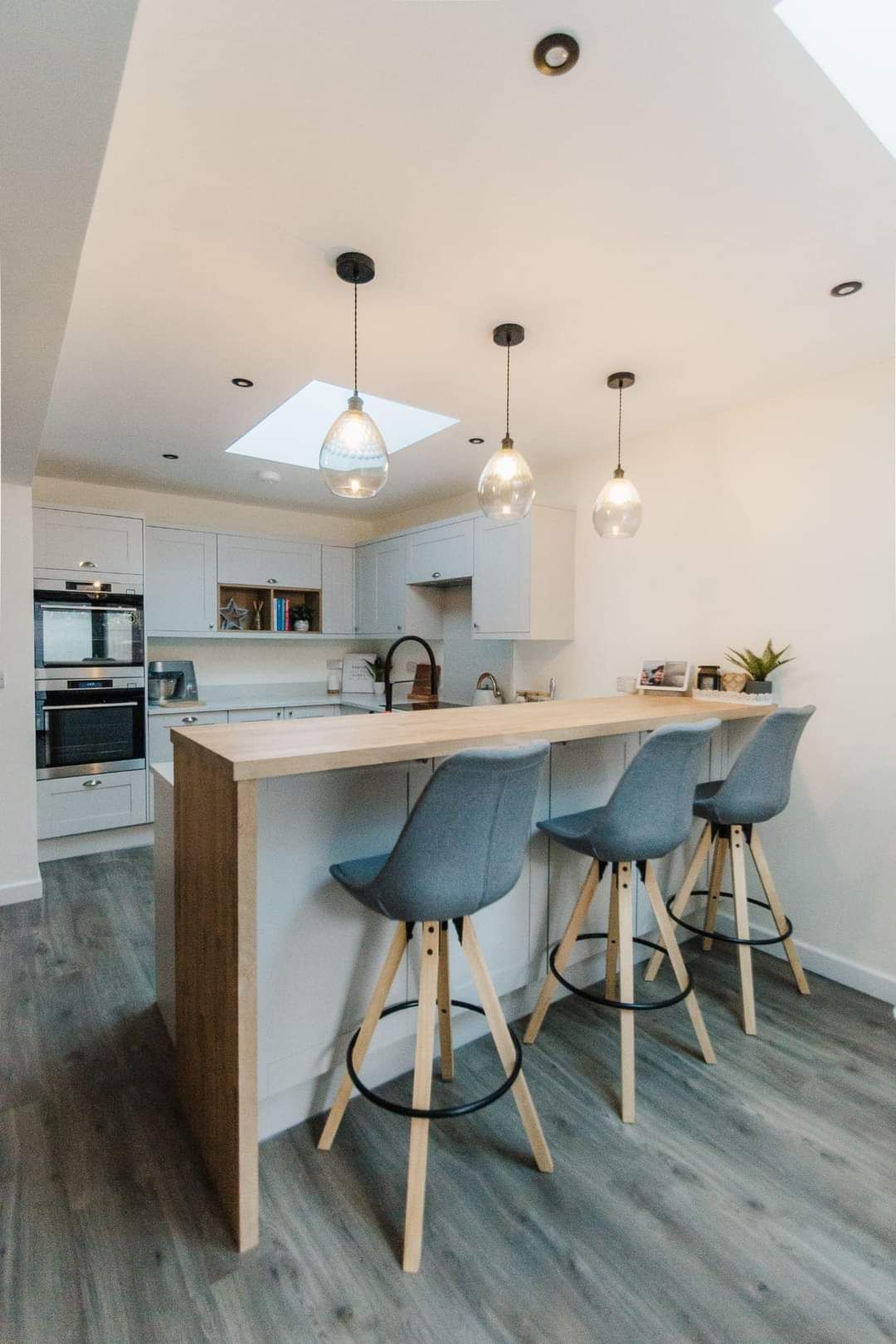 Kitchen with light wood breakfast bar, grey stools, white cabinets, and pendant lights.