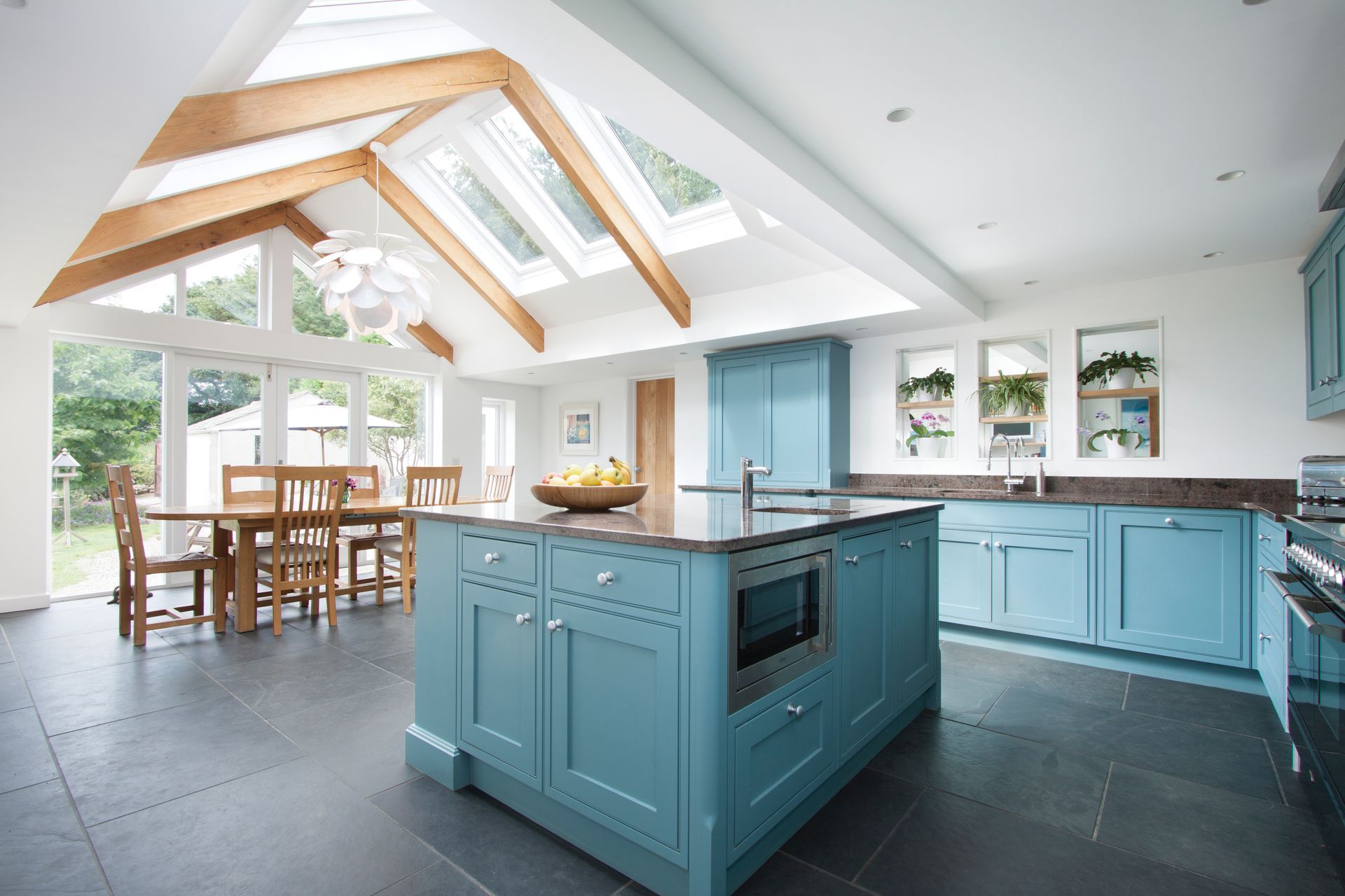 Spacious blue kitchen with island, skylights, and dining area. Black tile floor, wooden beams, and white walls.