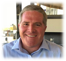 A smiling person with short hair, wearing a light blue button-down shirt, pictured against an indoor window background.