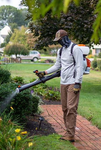 A person wearing a cap and mask uses a backpack sprayer to mist a garden bush near a brick pathway.