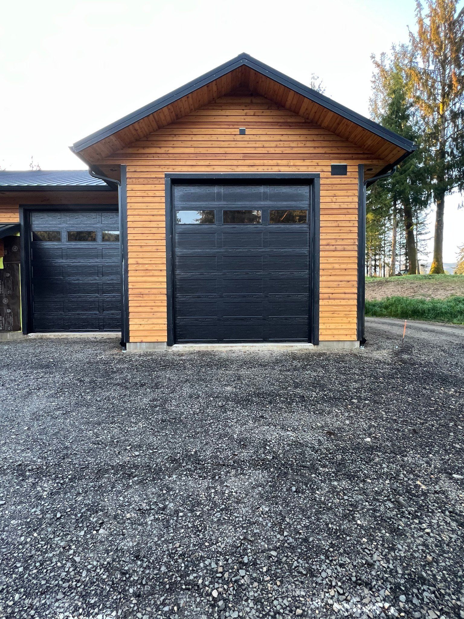 A wooden garage with a black garage door is sitting on top of a gravel driveway.