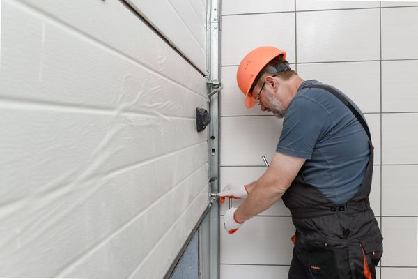 Man in orange hard hat and overalls repairing white garage door.