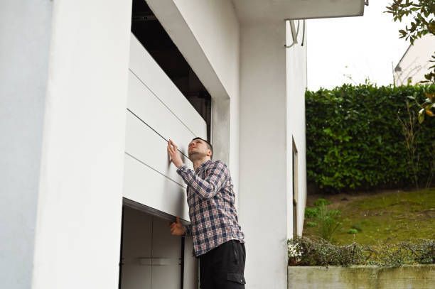 Person lifts garage door manually outside white building with greenery in the background.