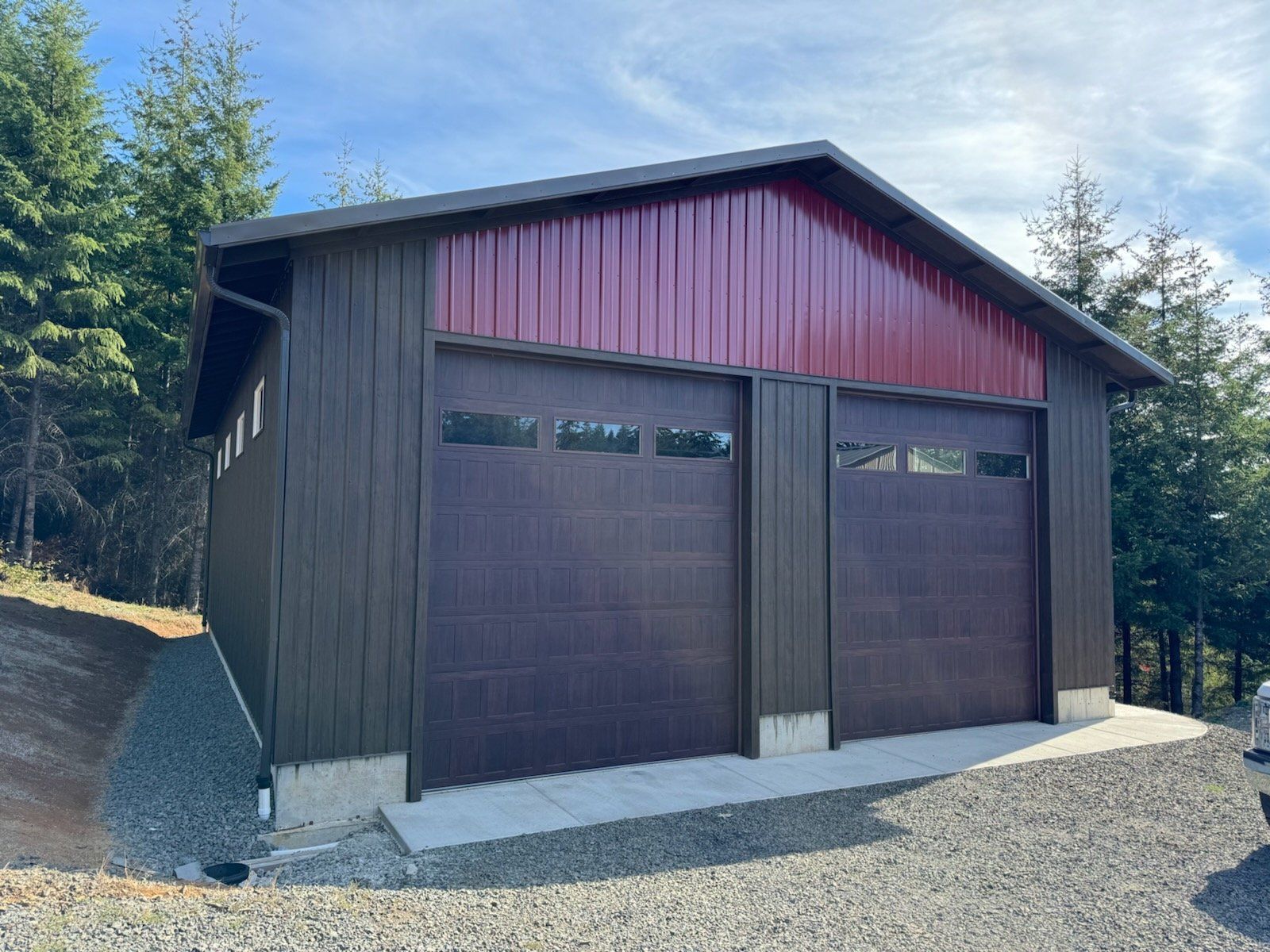 A garage with three garage doors and a red roof is sitting on top of a gravel hill.
