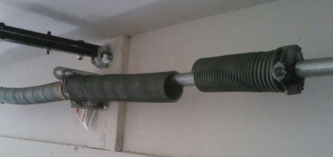 Garage door spring mechanism: a grey spring coils around a silver rod, connected to mounting hardware. The mechanism is near the ceiling.