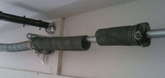 Garage door spring mechanism: a grey spring coils around a silver rod, connected to mounting hardware. The mechanism is near the ceiling.