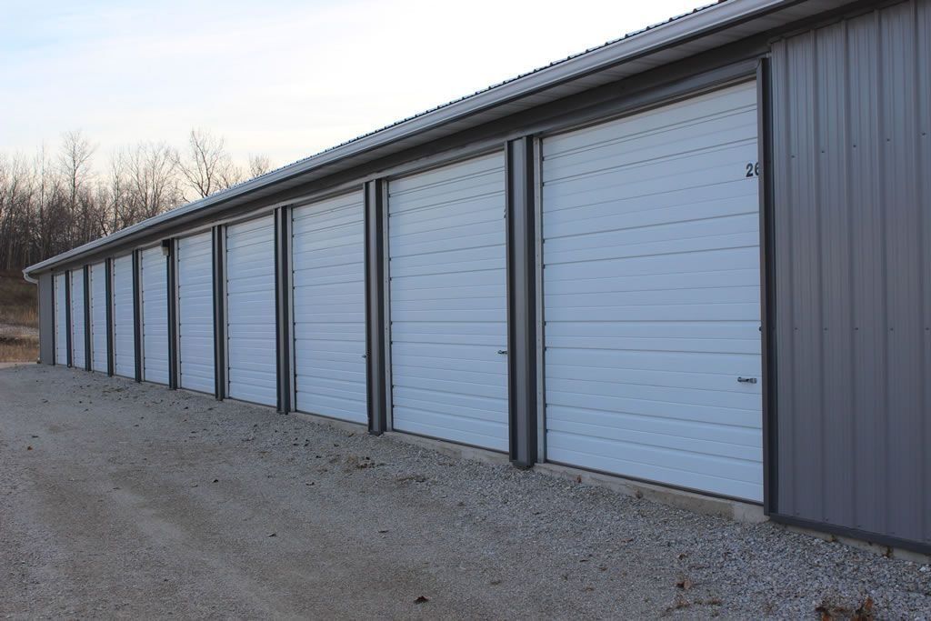 Row of white storage unit doors under a gray metal roof, gravel driveway.
