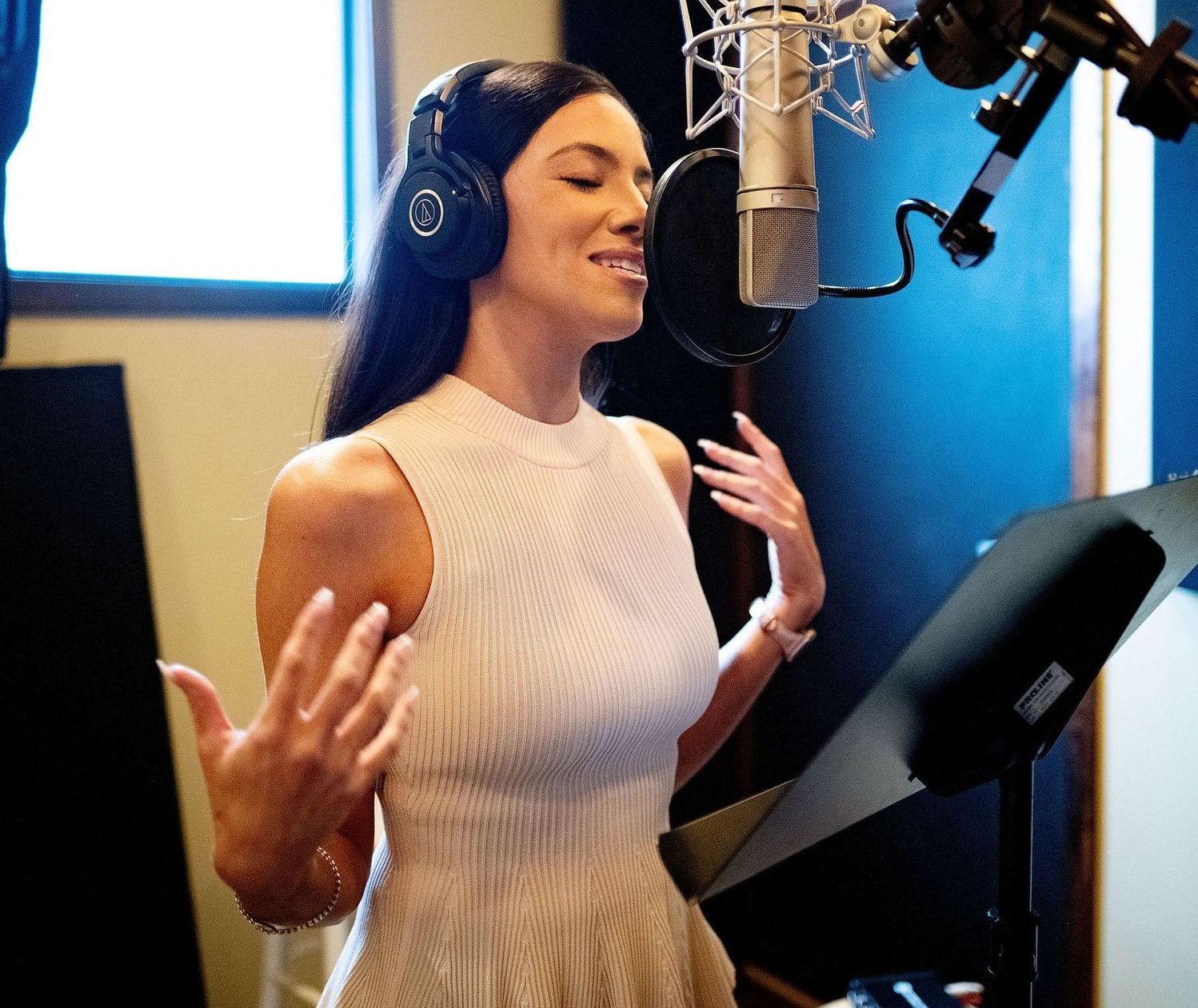 Iris voicing into a microphone in a recording studio, wearing headphones and a cream dress.
