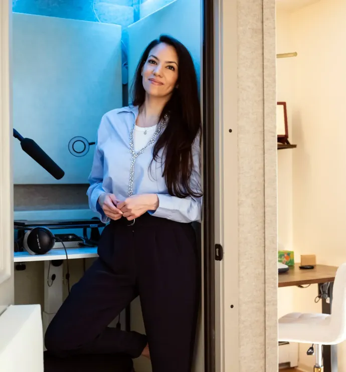Iris in light blue shirt and dark pants leans in doorway. Interior of recording booth visible behind her.