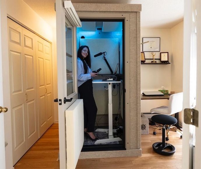 Iris in a soundproof booth, working at a standing desk with a microphone. Office setting with wood floors.