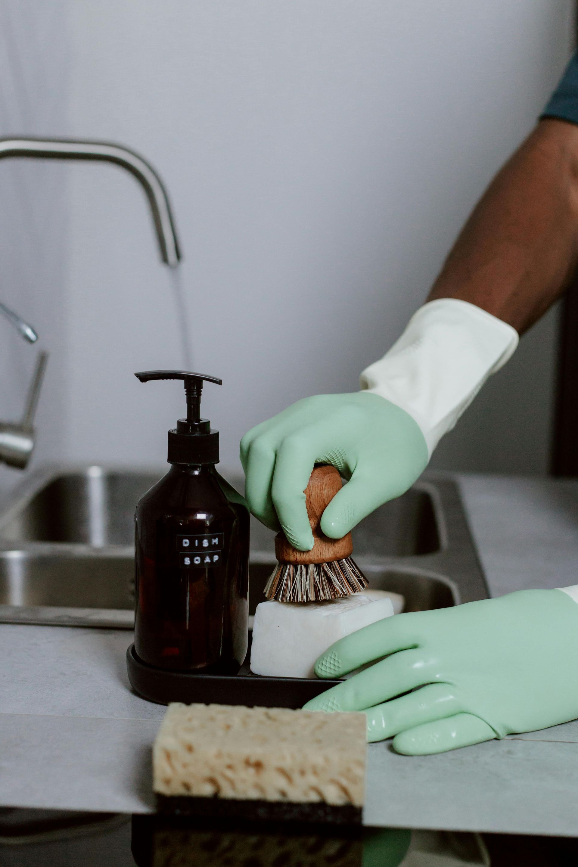 Person in green gloves uses a brush and soap to wash dishes in a kitchen sink.