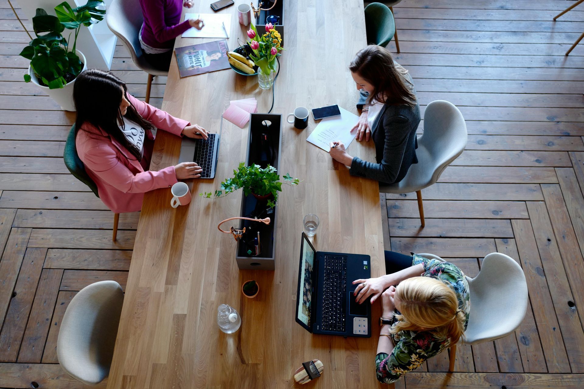 People working at a long wooden table in an office. Two are using laptops, one is writing.