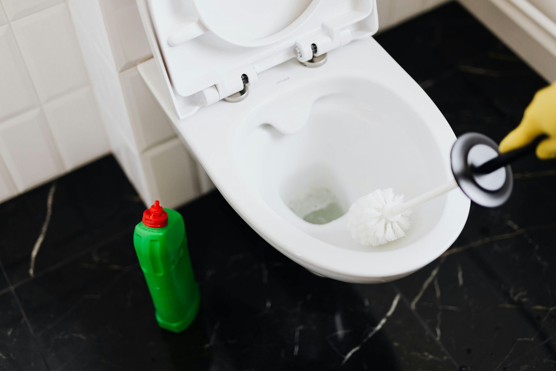 A person wearing yellow gloves cleans a white toilet with a brush, green cleaner on the floor.