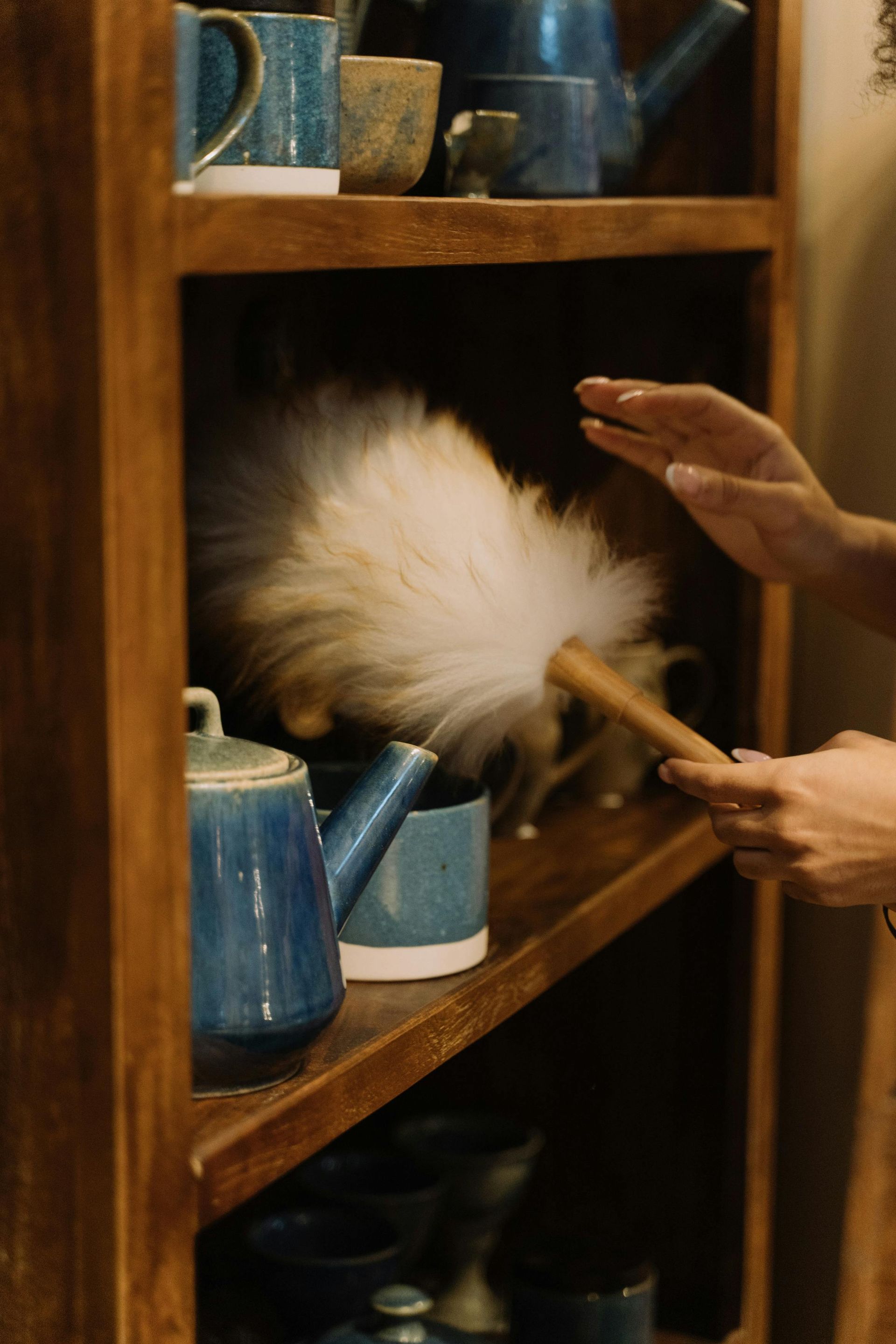 Person dusting a wooden shelf with blue ceramic teacups and mugs using a fluffy duster.