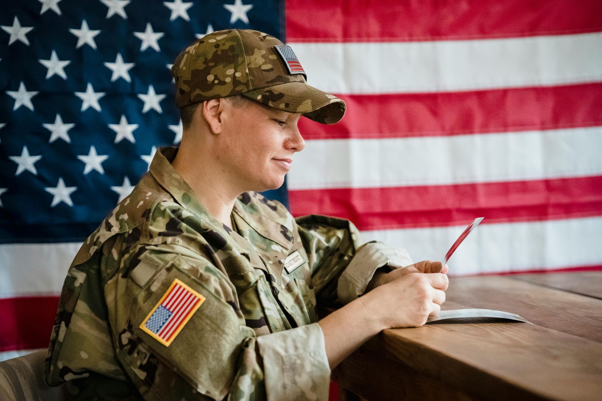 U.S. soldier in camouflage writing at a desk in front of an American flag