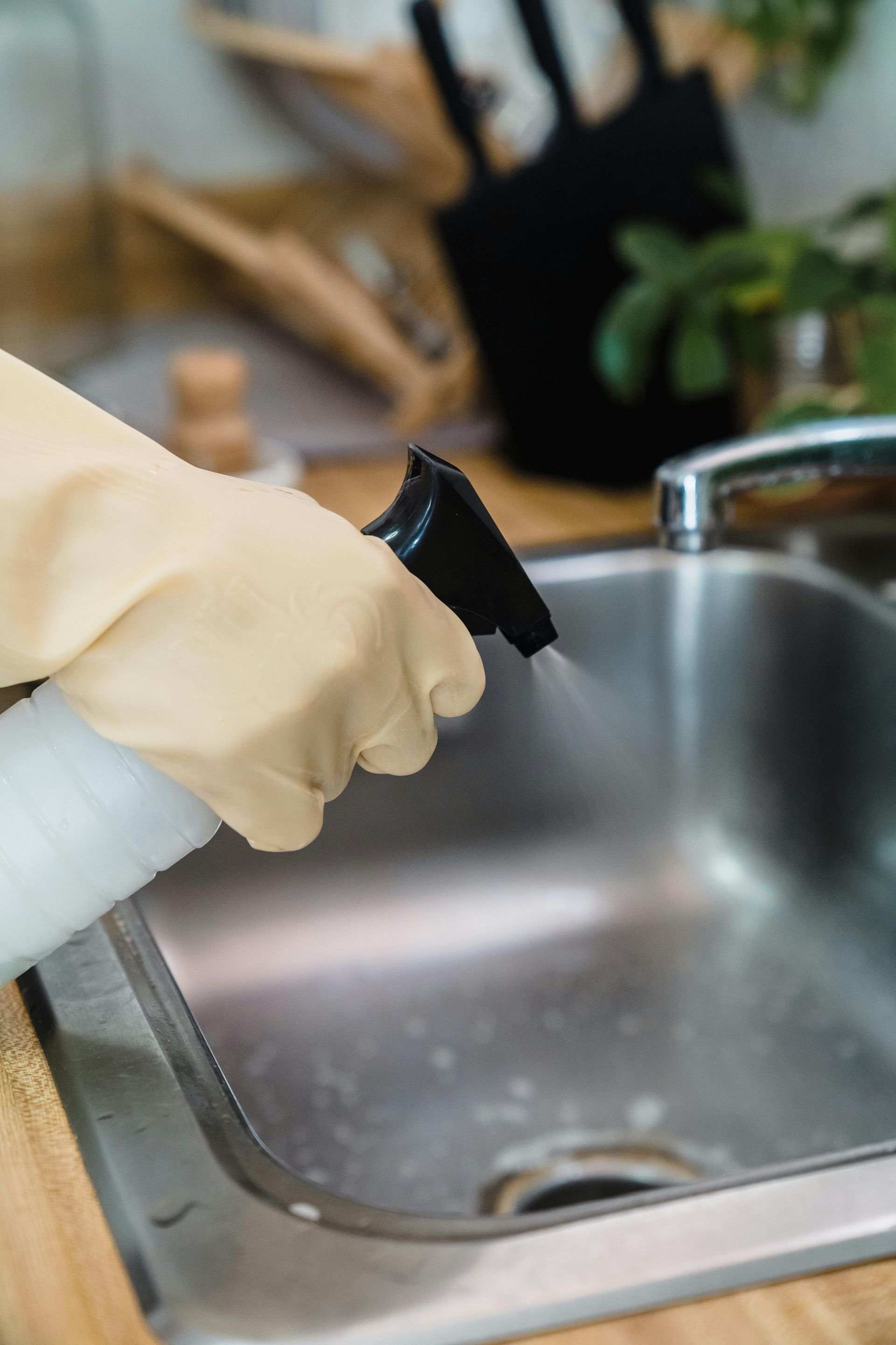 Person wearing yellow gloves spraying cleaner into a stainless steel sink.
