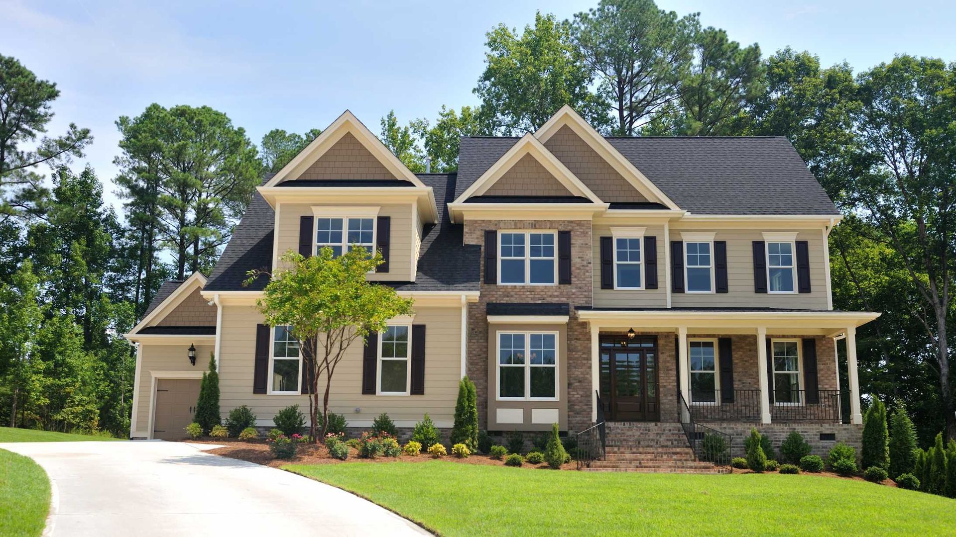 Two-story house with tan siding, brick accents, black shutters, and a lush green lawn, with trees in the background.