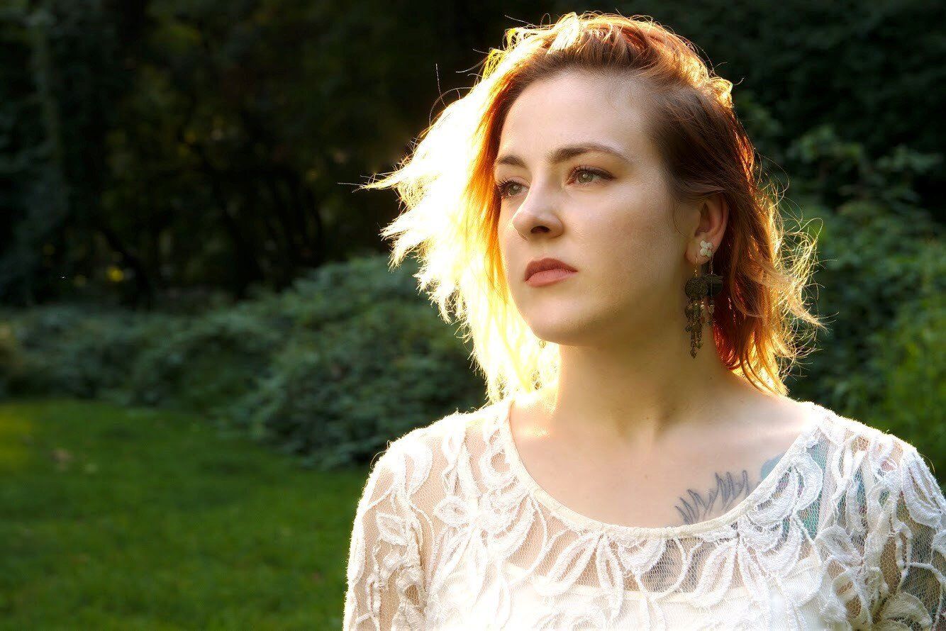 A woman in a white lace top with a tattoo on her chest is standing in the grass.