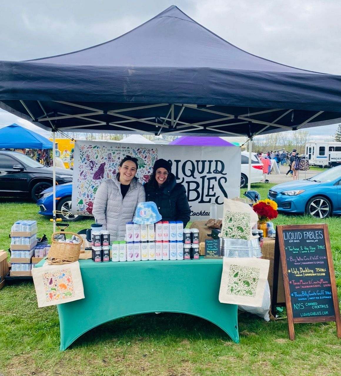 Two women are standing in front of a table under a tent that says liquid bliss.