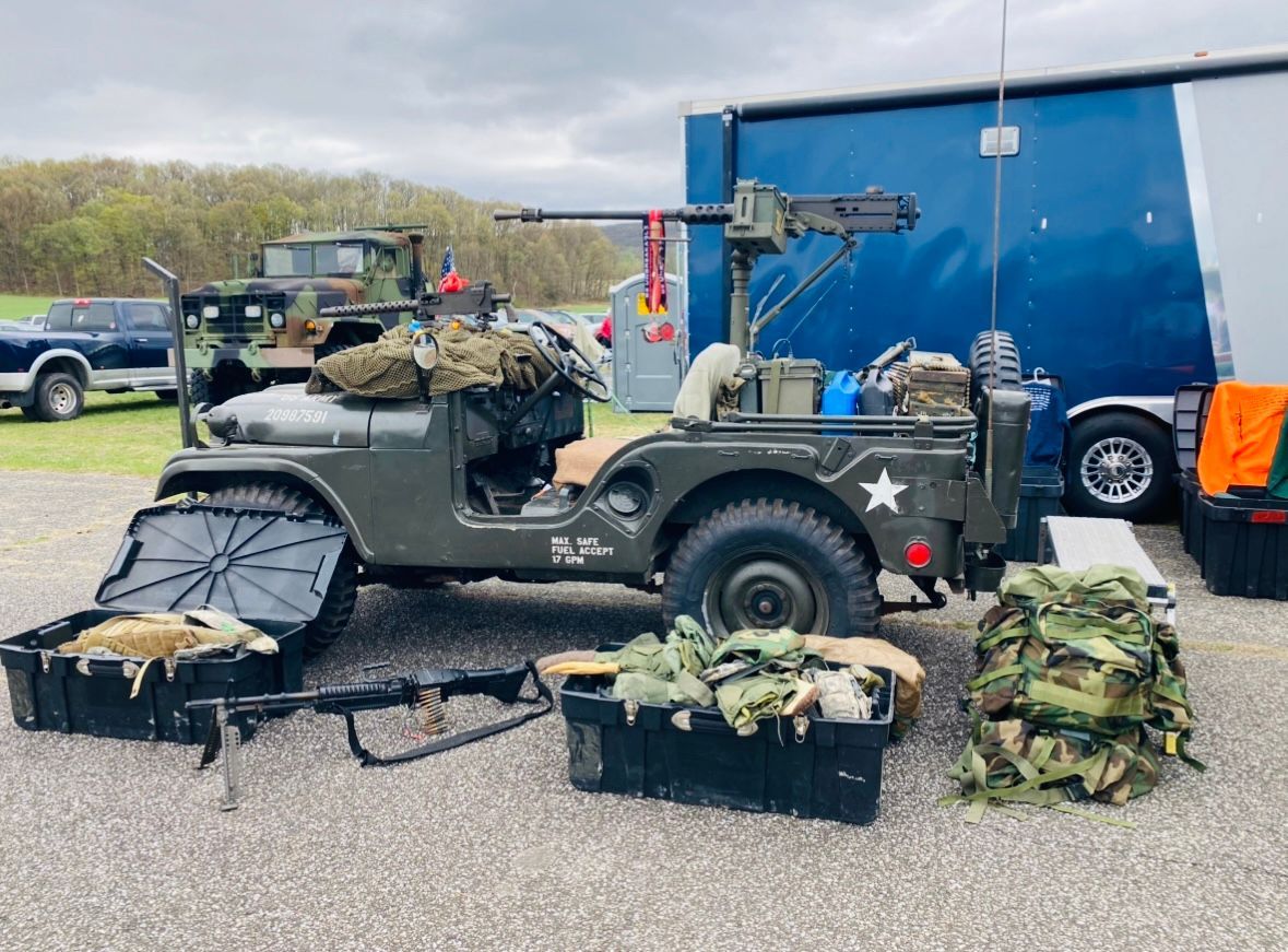 An old military jeep is parked in a gravel lot next to a trailer.