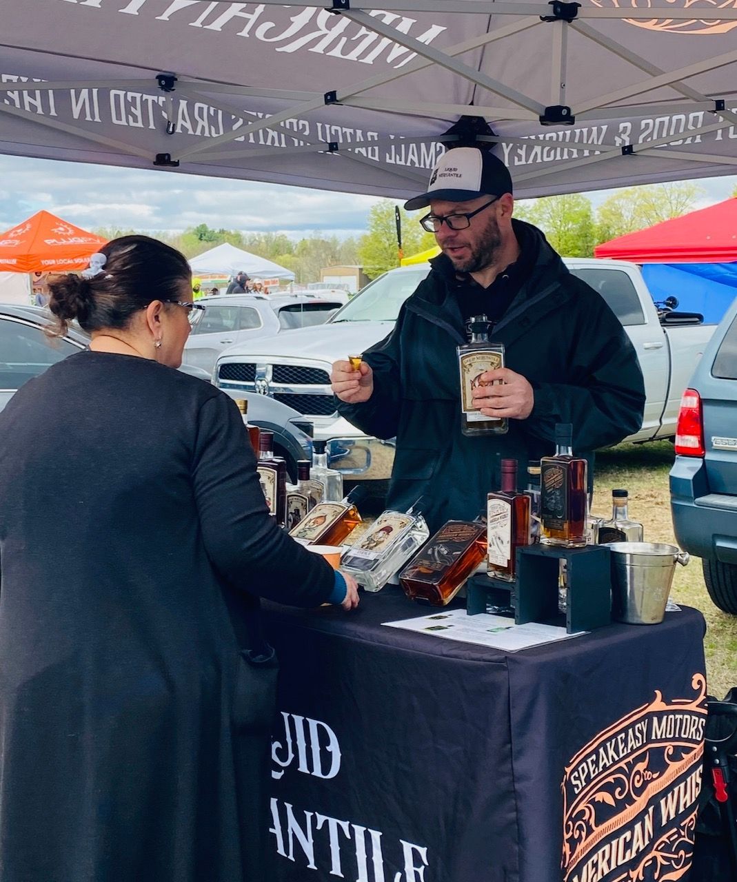 A man and a woman are standing at a table with bottles of alcohol on it.