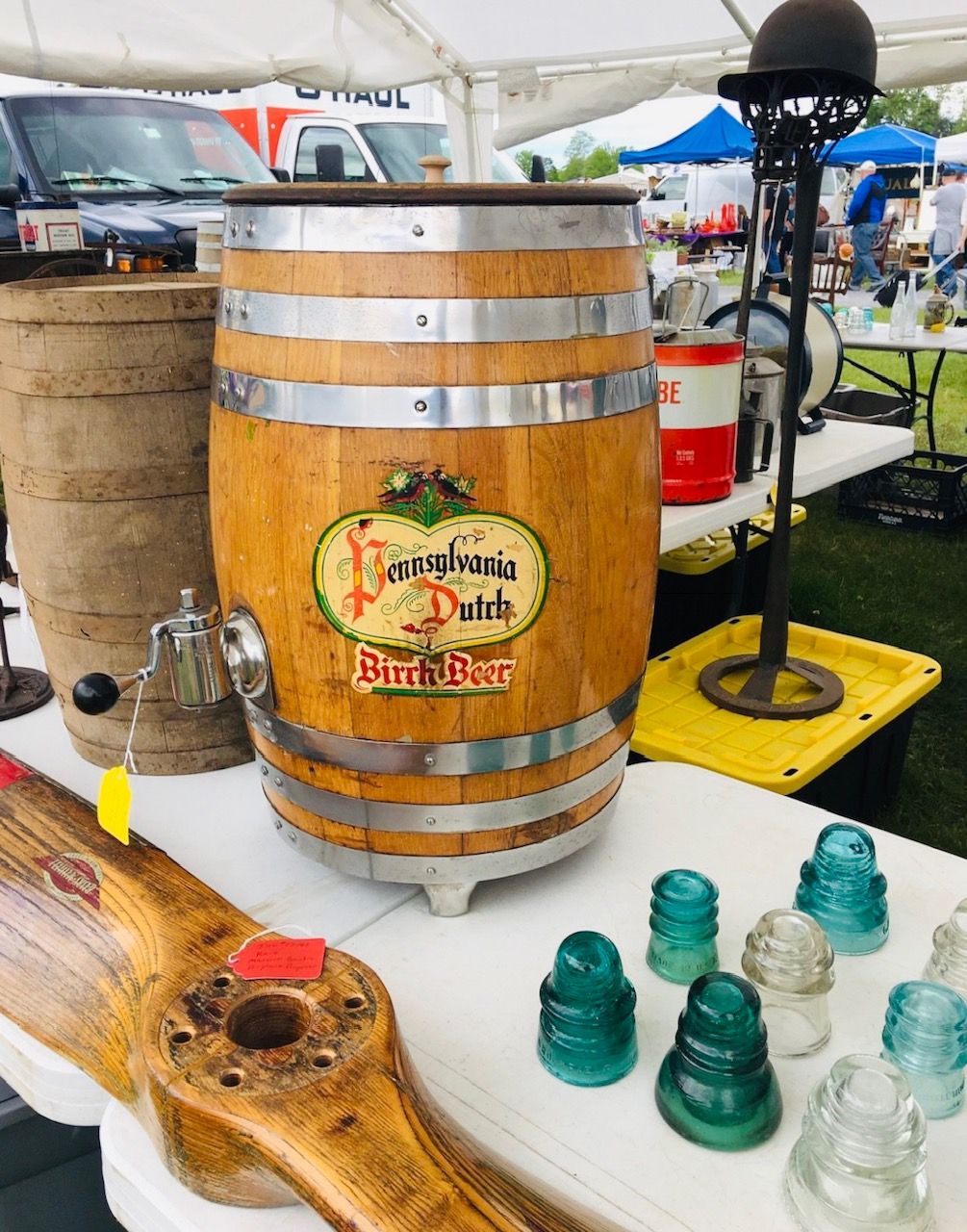 Wooden keg with Pennsylvania Dutch Birch Beer label at outdoor market, surrounded by various antiques.