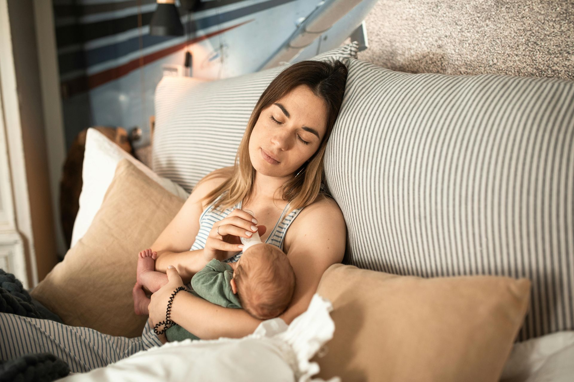 Woman sitting in bed, bottle-feeding infant. Beige pillows, striped blanket; soft lighting.