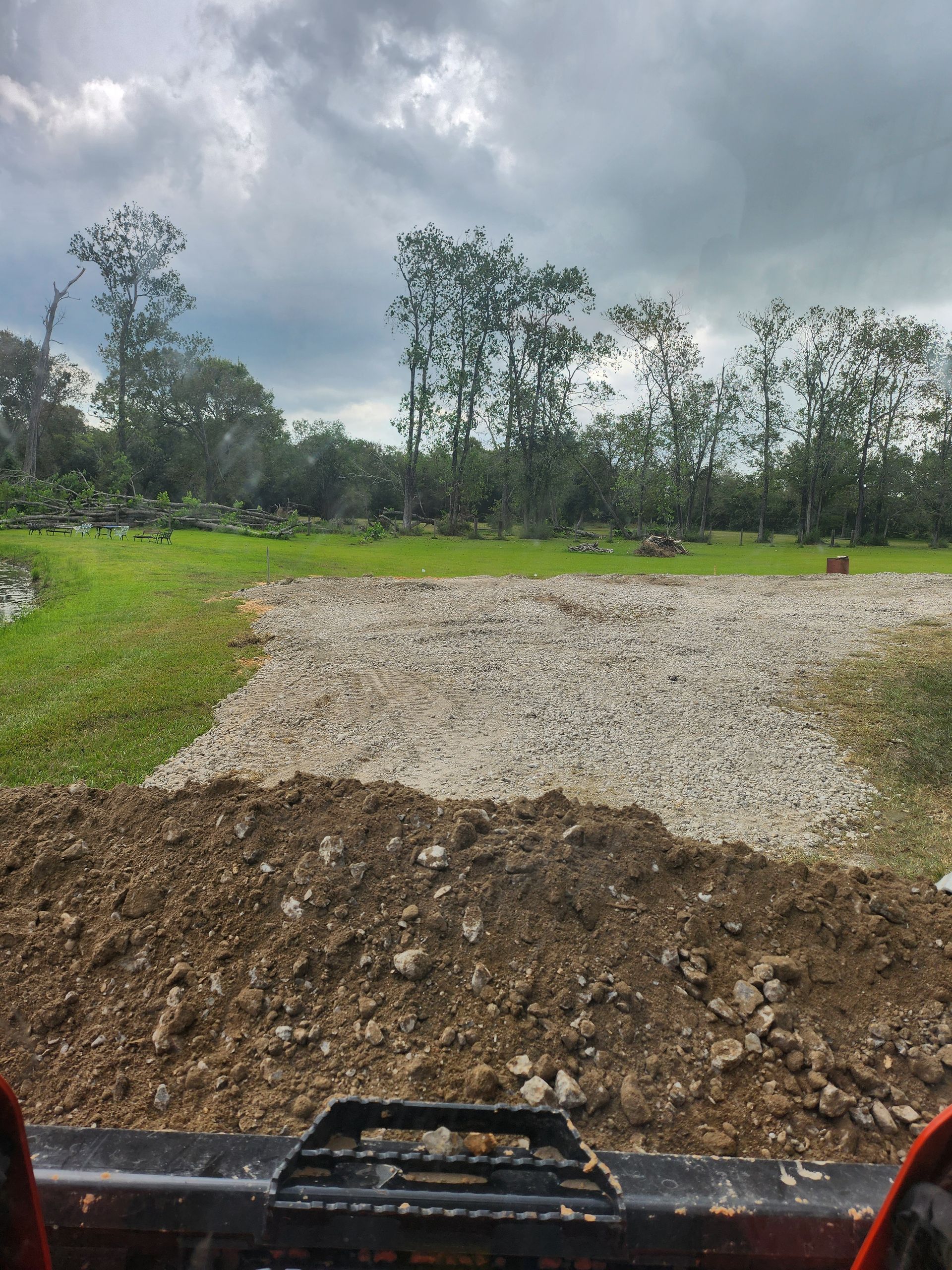 A view from a construction vehicle: a pile of dirt in the foreground, gravel area in the middle, and trees under a cloudy sky.
