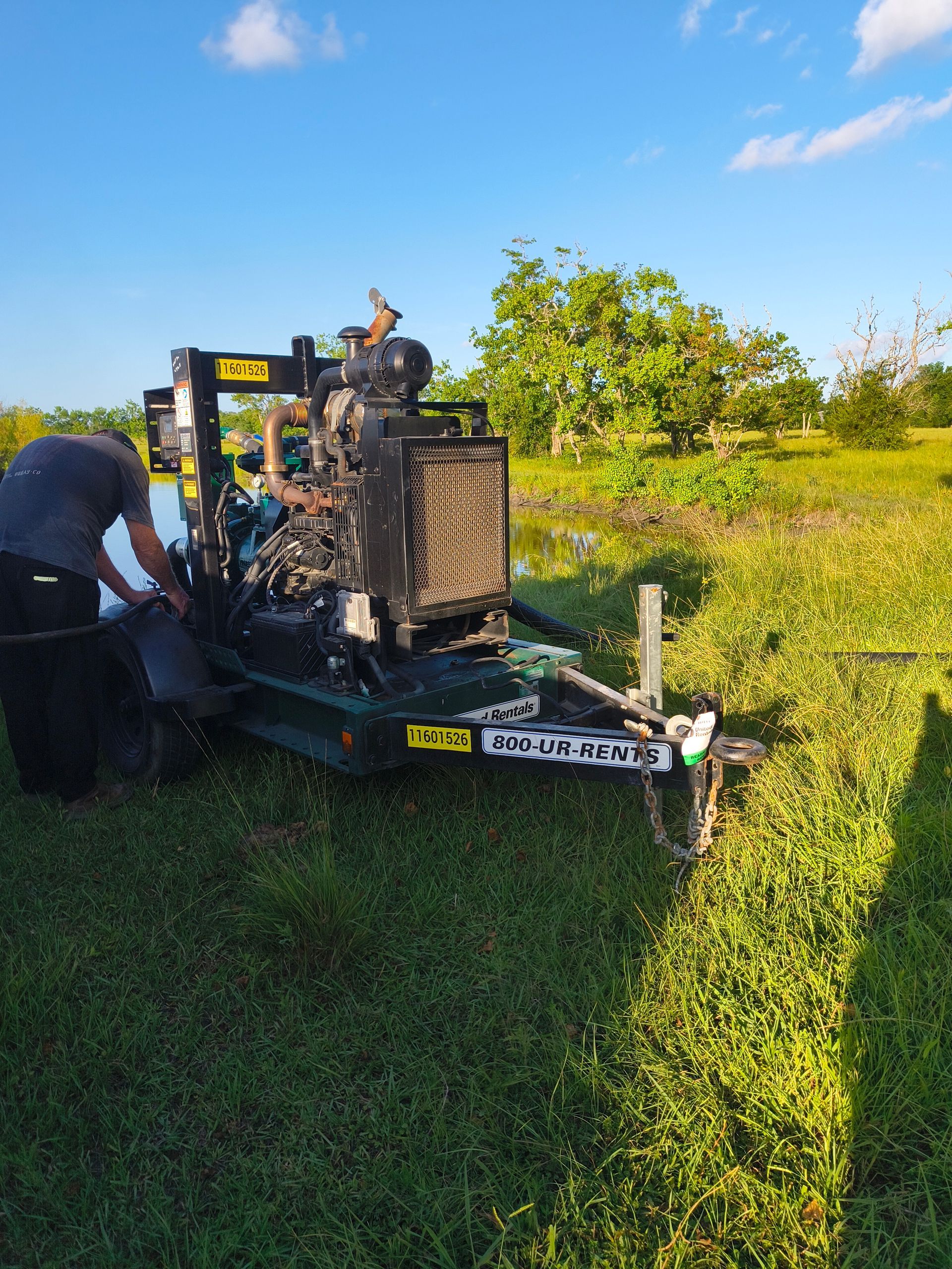 A man near a green industrial pump on grass, next to a flooded area with trees under a blue sky.