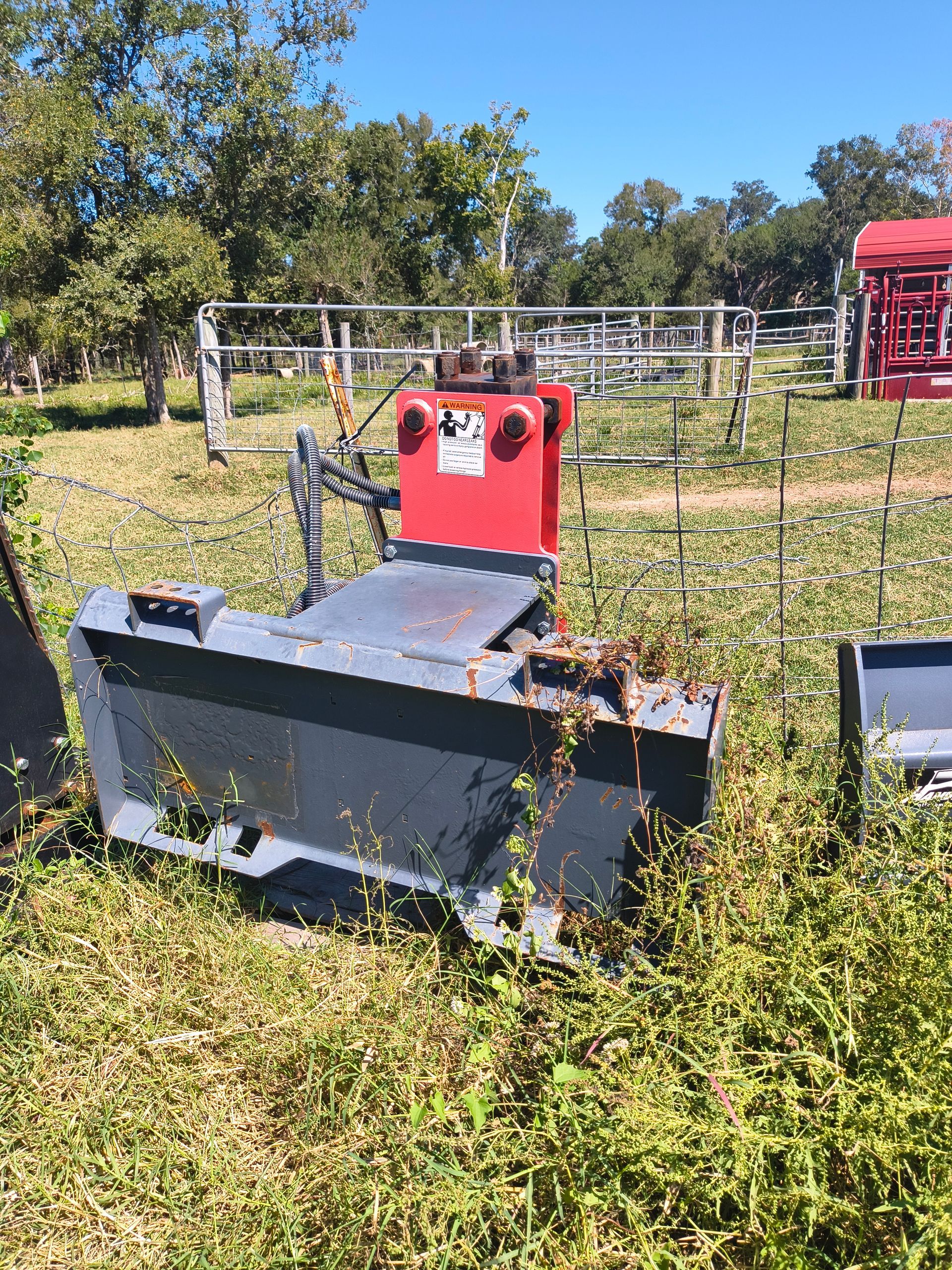 A red and gray tractor attachment with a blade, sitting in a grassy field on a sunny day.