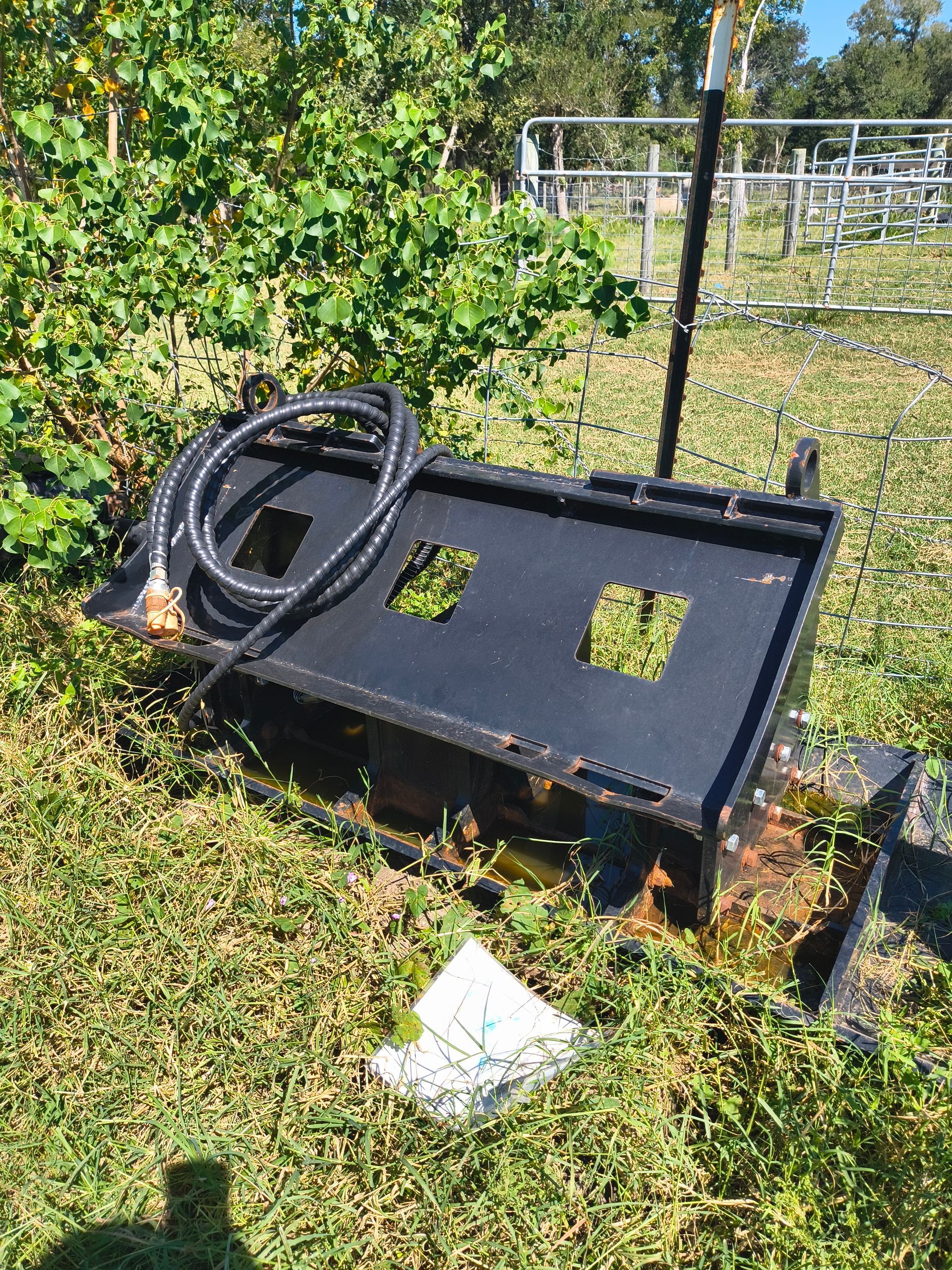 Black metal skid steer attachment on grass, with hose and handle. In outdoor setting.