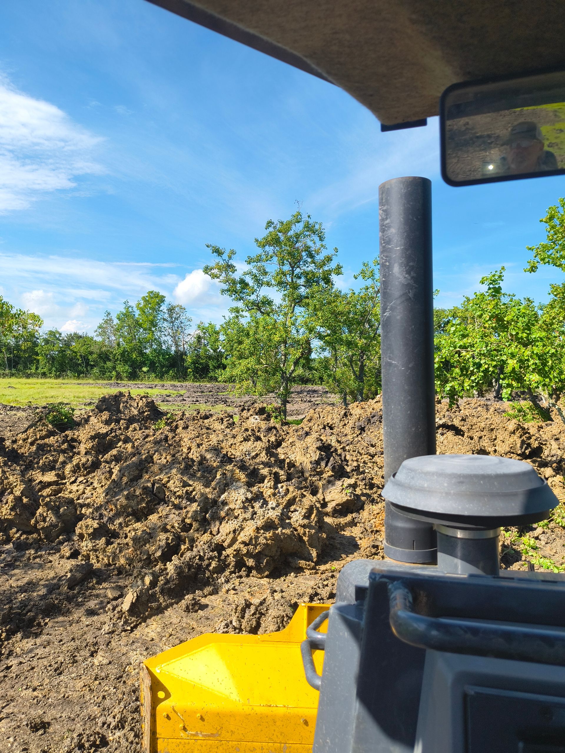 View from a bulldozer's cab, grading earth. Yellow blade, black exhaust, blue sky, trees in the background.