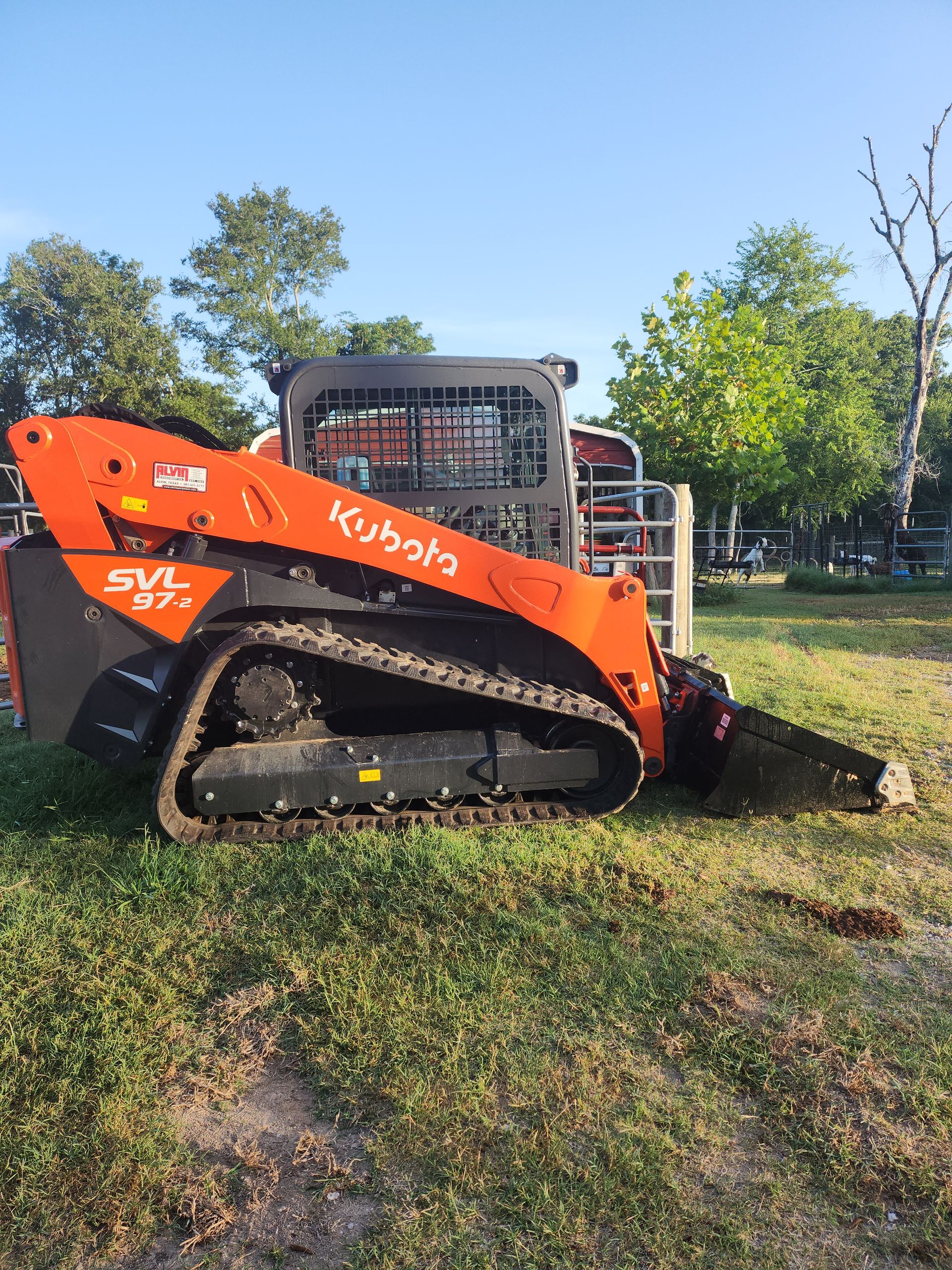 Orange Kubota SVL75-2 compact track loader on grass, next to a fence under a blue sky.