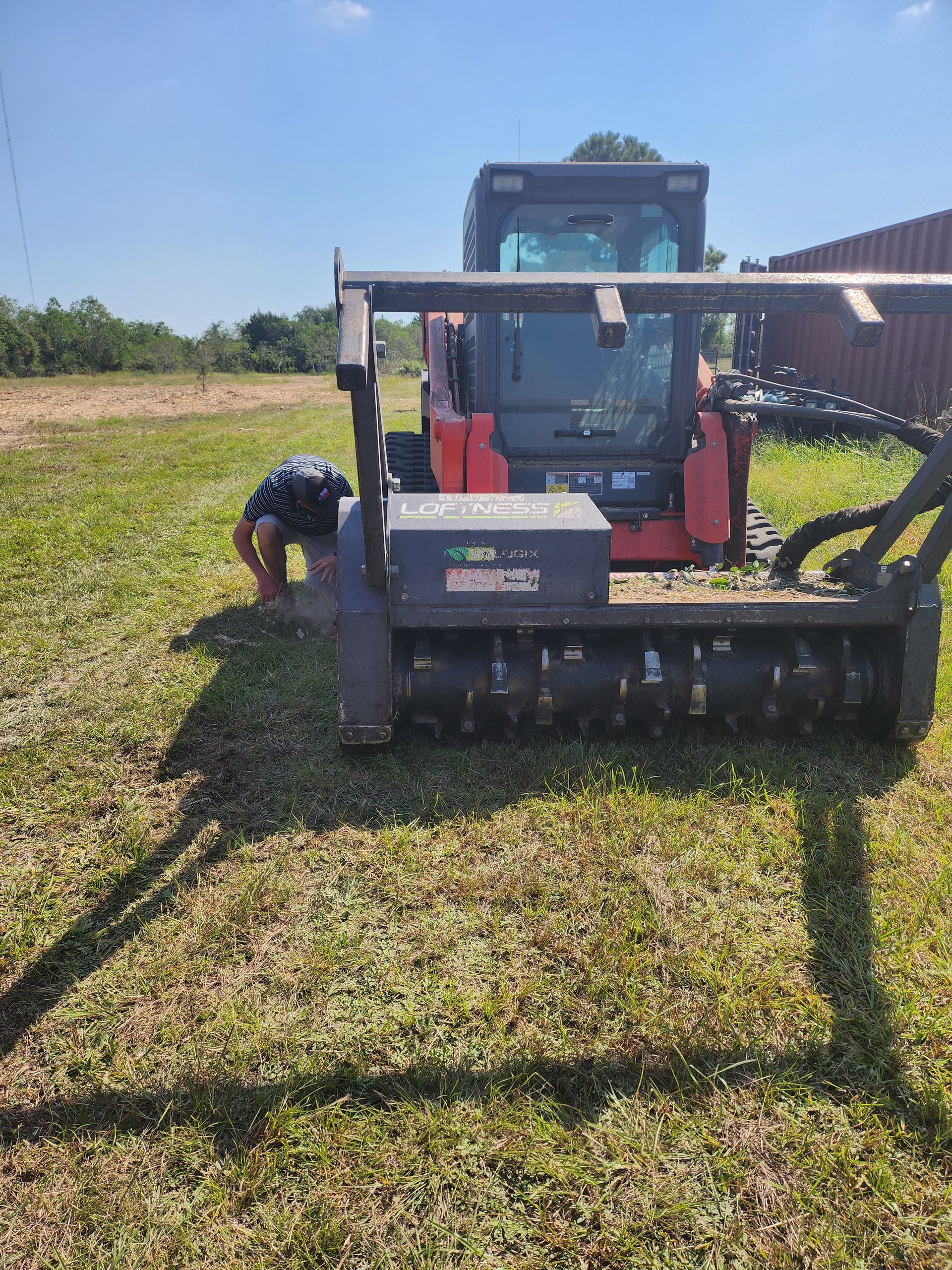 Person near a skid steer with brush cutter in a grassy field on a sunny day.
