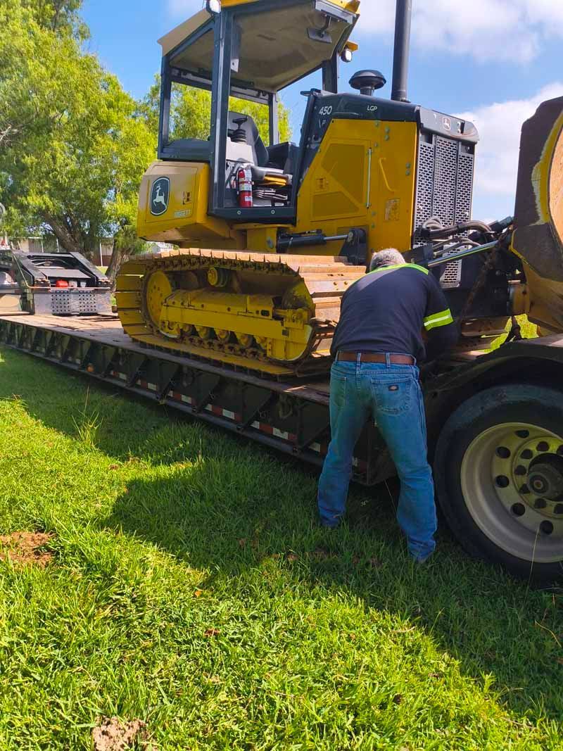 Man working on a yellow bulldozer on a trailer, sunny outdoor setting.