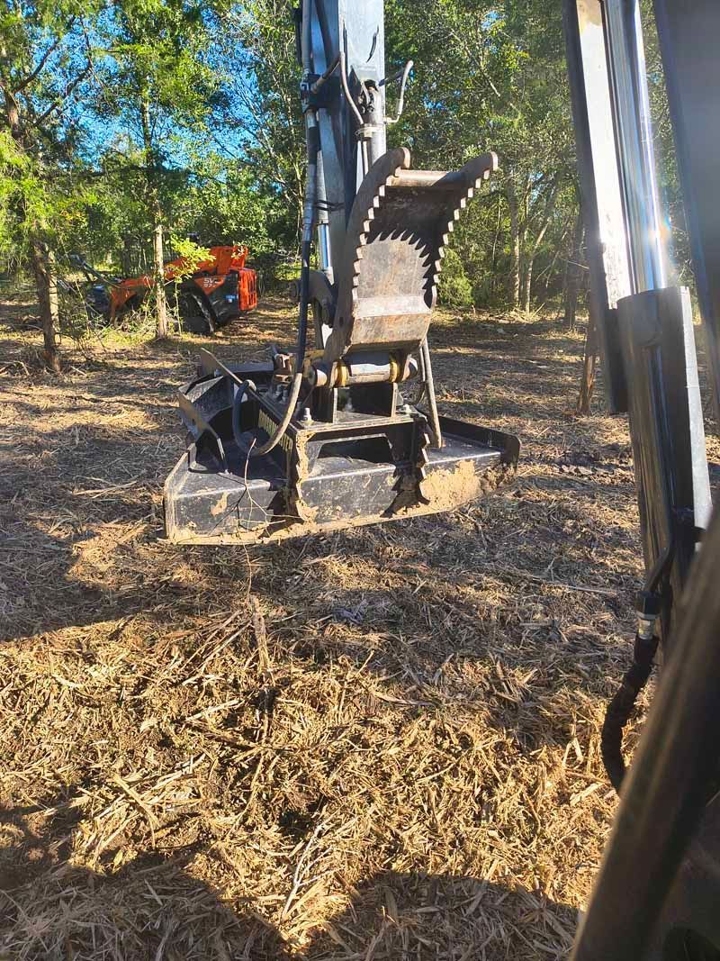 Excavator with saw attachment clearing debris in a wooded area. Orange machine in the background.