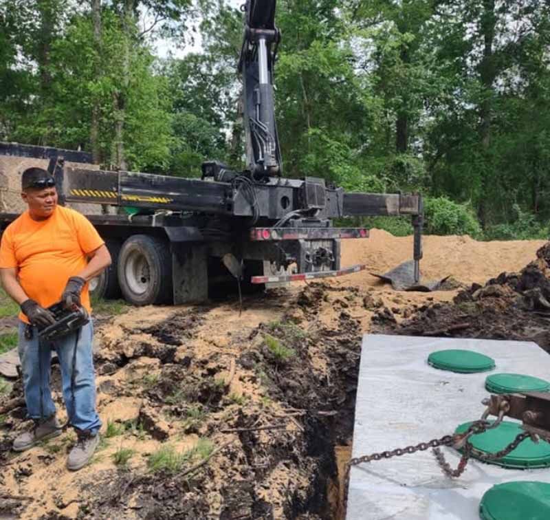 Man operating crane, installing septic tank. Orange shirt, blue jeans, dirt, green lids.