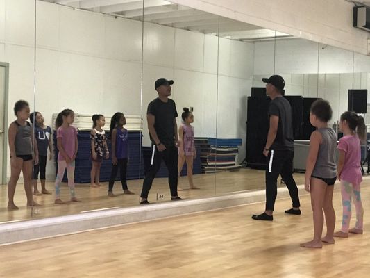 A group of children are standing in front of a large mirror in a dance studio.