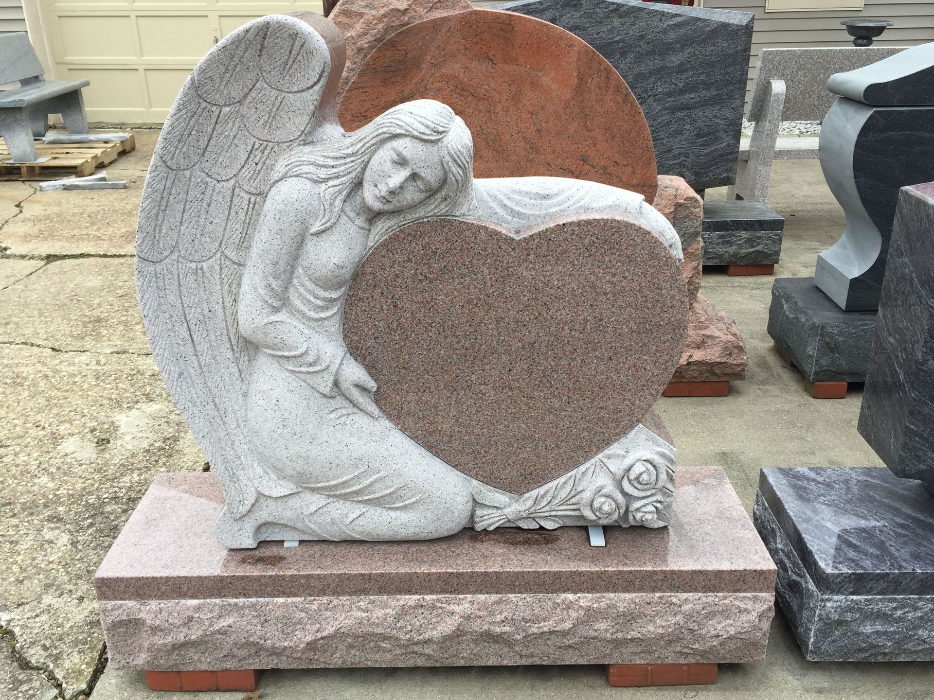 Angel sculpture embracing a heart-shaped memorial. Granite stone, red-toned, outdoors.