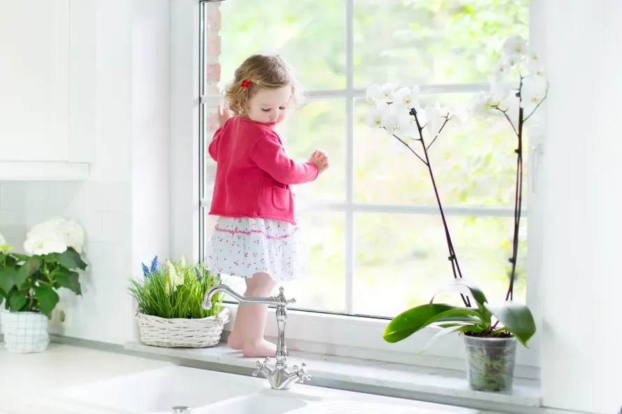 A little girl is standing on a window sill in a kitchen.