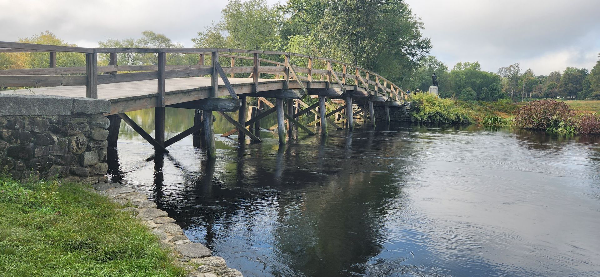 Wooden bridge over dark water in a park setting. Overcast sky, greenery on banks.