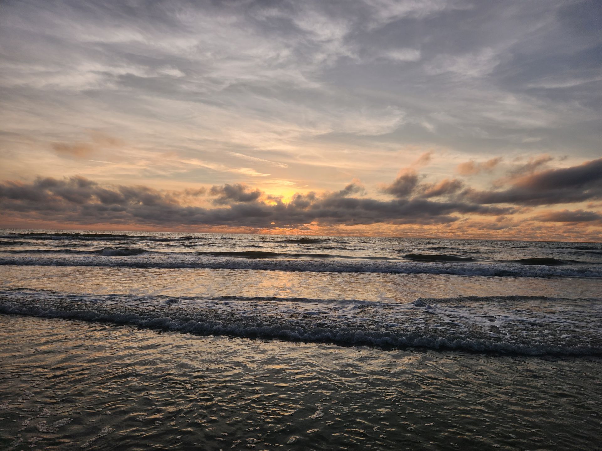 Sunset over the ocean, waves lapping the shore. Cloudy sky with golden and grey hues.