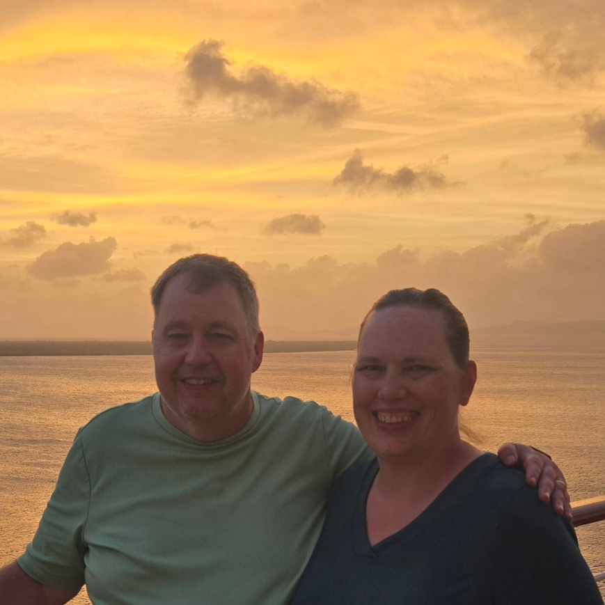 Couple smiling, posing together outdoors at sunset, with ocean in background.