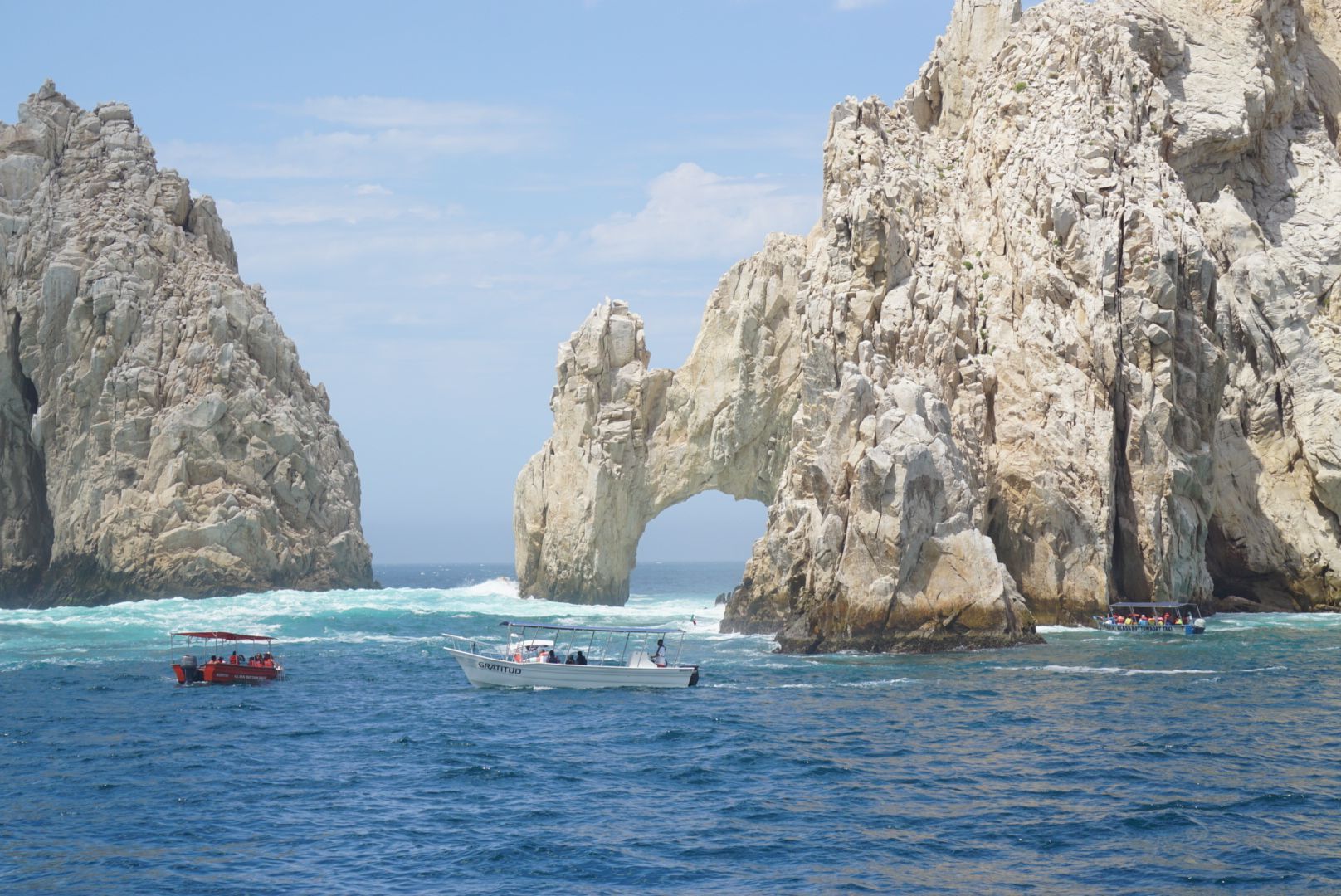 Rocky cliffs arch over blue ocean with boats in Cabo San Lucas.
